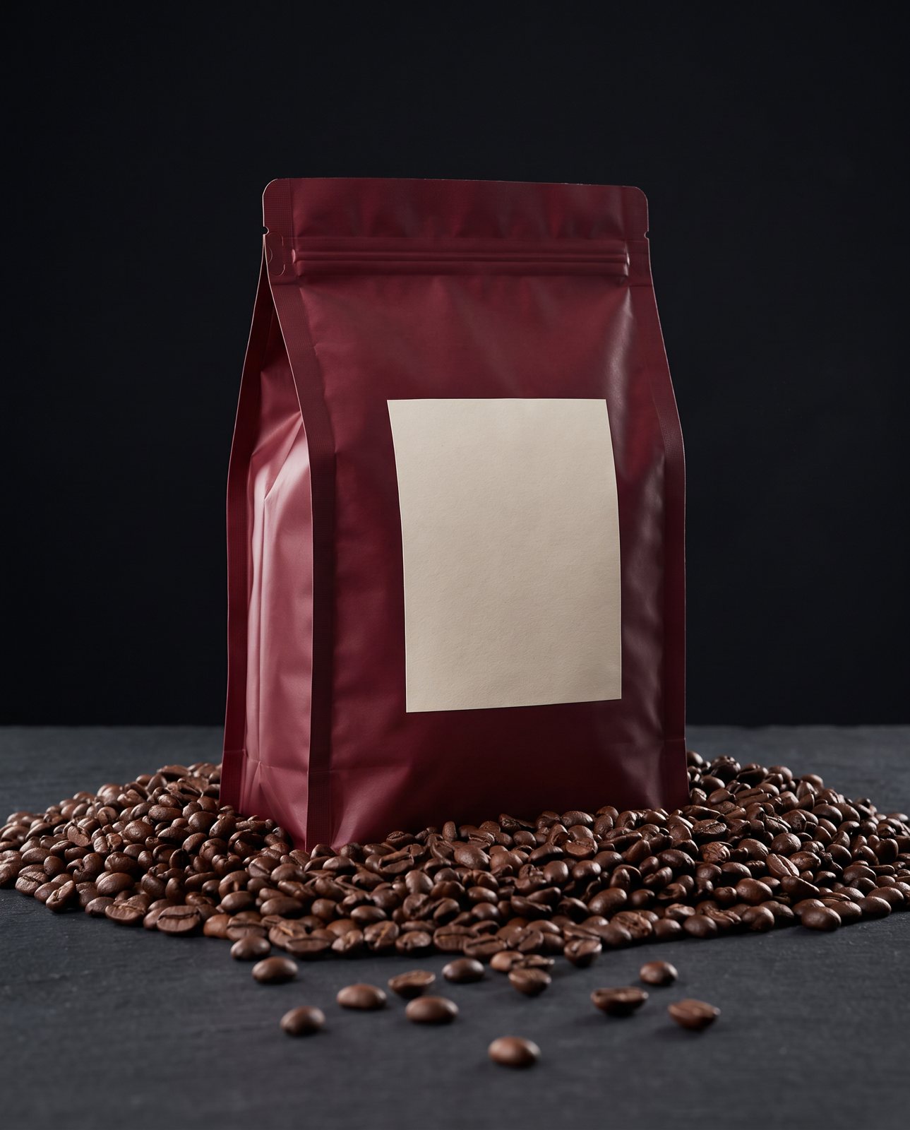 The burgundy coffee pouch with its blank cream label standing in a heap of coffee beans against a black studio backdrop