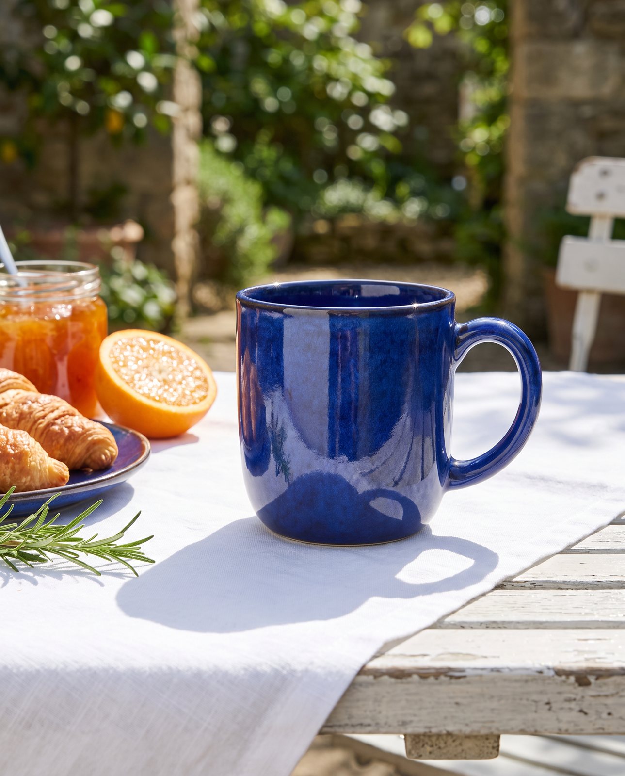 The cobalt blue glazed mug on a white-clothed garden table with croissants and orange halves in sunlight