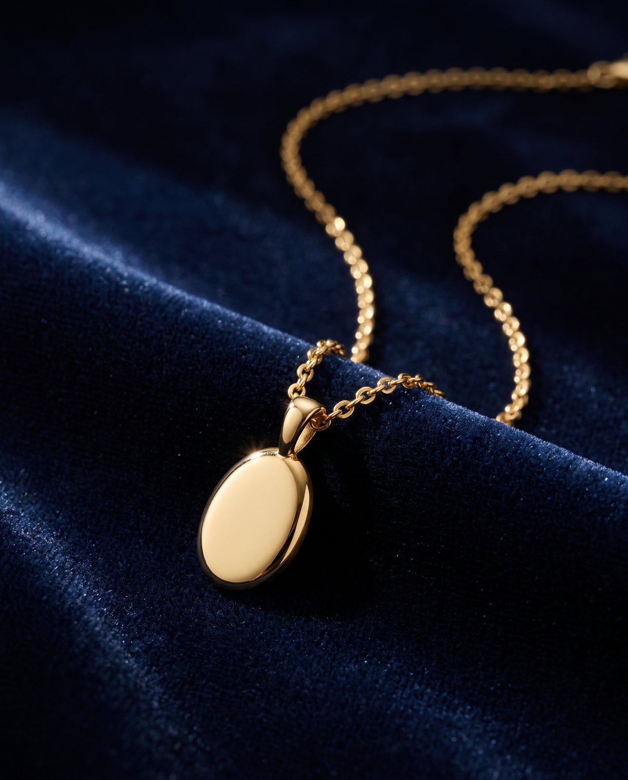 A close-up of a small gold oval pendant on a fine chain, draped over midnight blue velvet