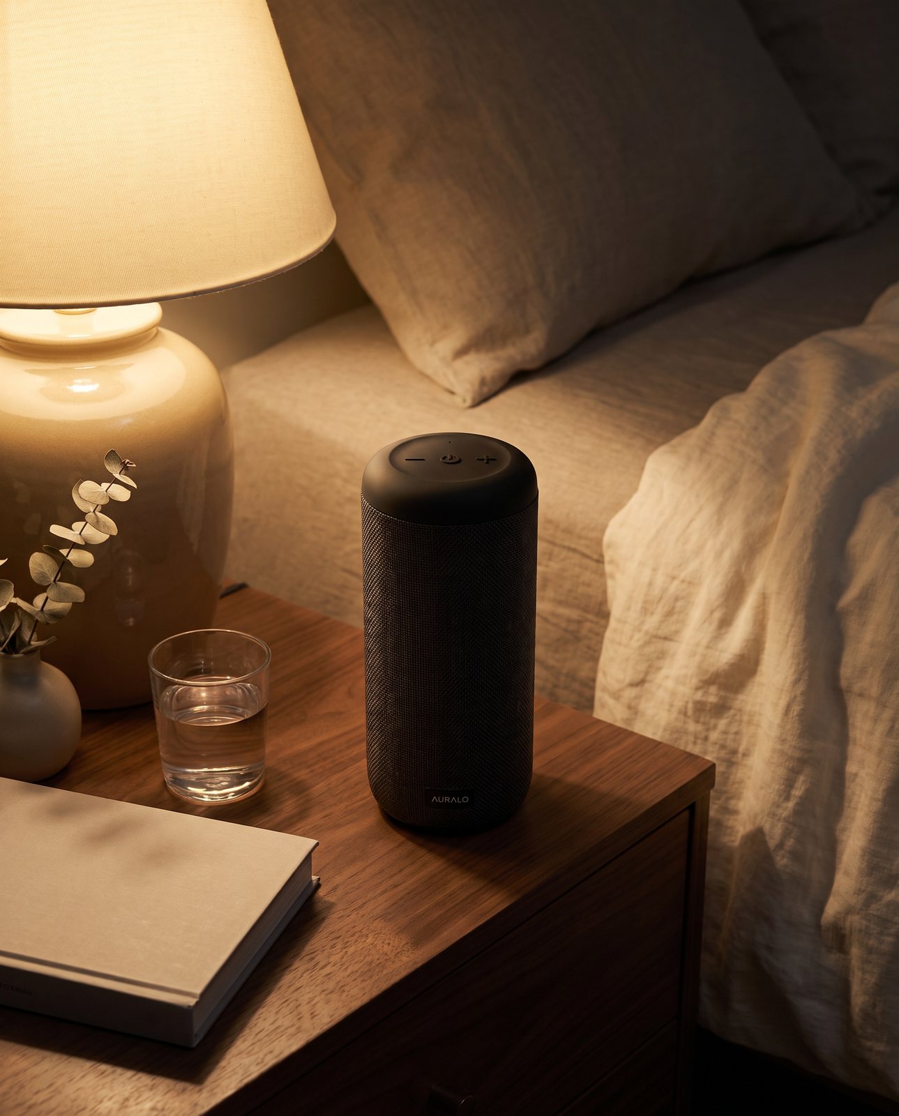 The same speaker on a walnut bedside table at night beside a glass of water, a book and a lit lamp